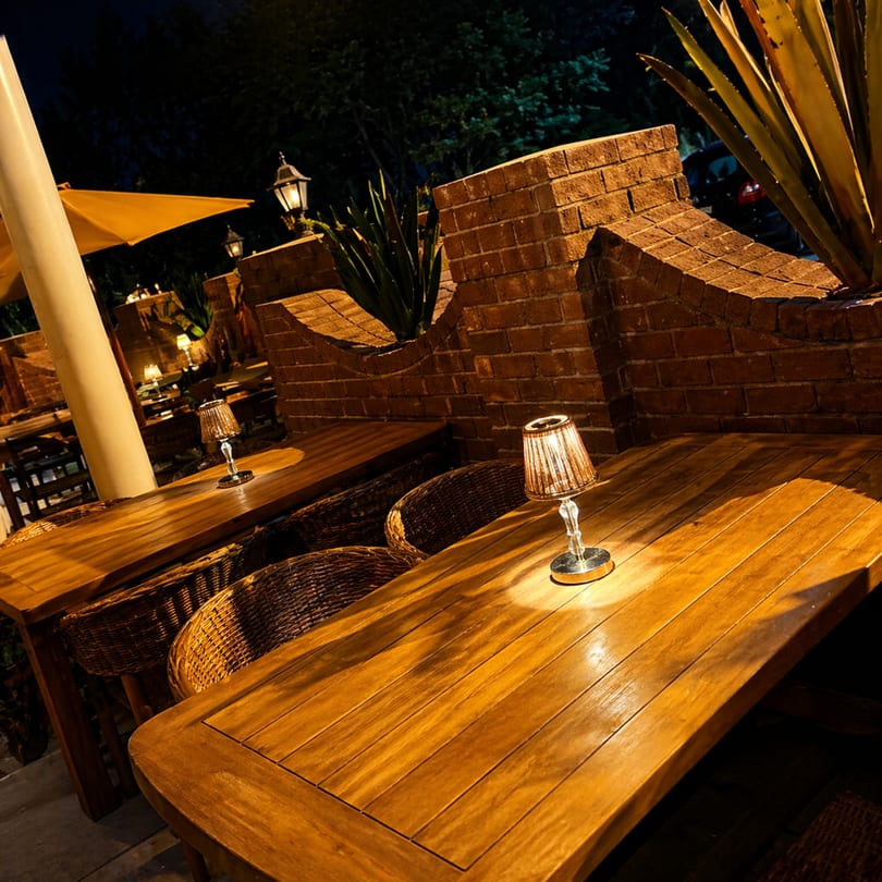 Outdoor restaurant patio at night with wooden tables, wicker chairs, and small lamps illuminating the space.