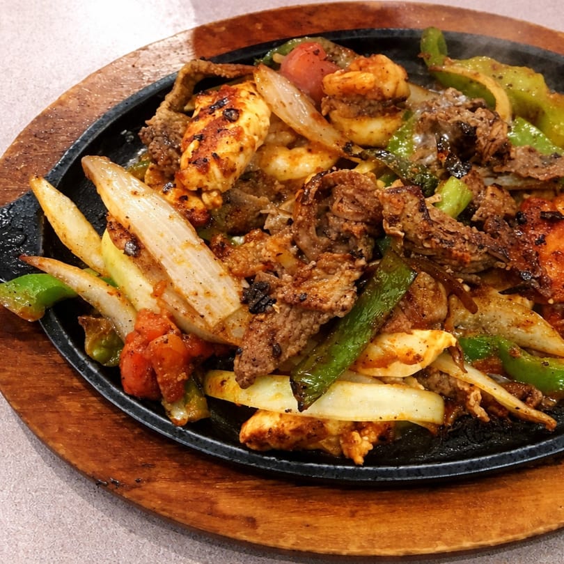 Sizzling fajitas with steak, chicken, shrimp, onions, and bell peppers on a cast iron platter.
