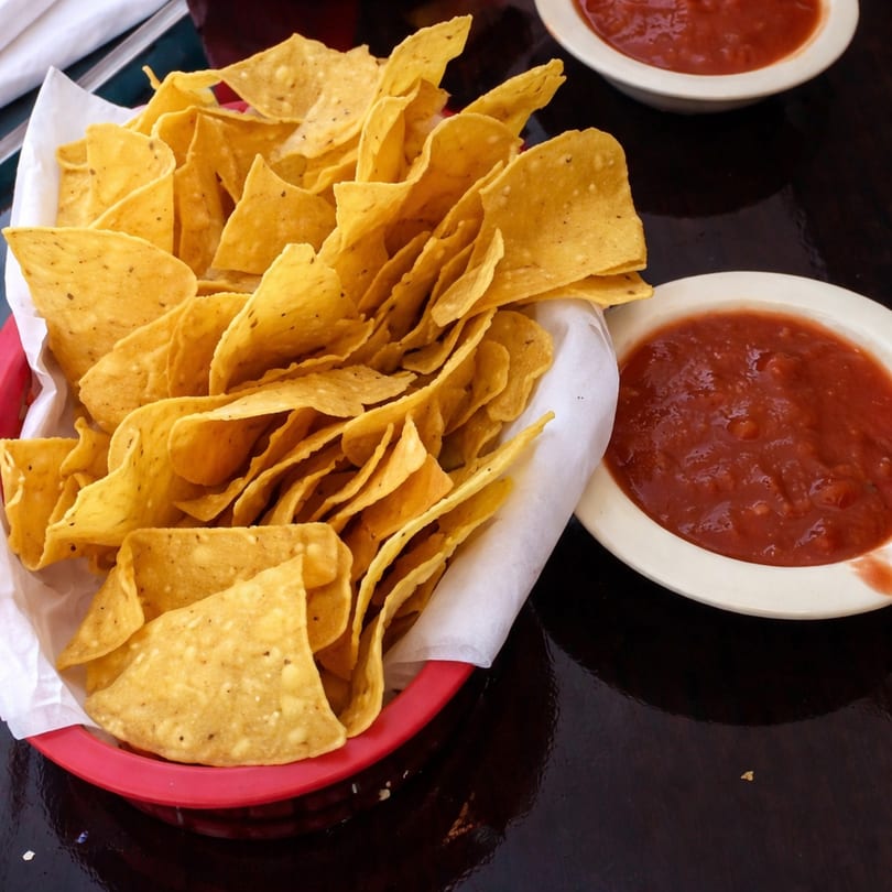 A red basket filled with tortilla chips and two bowls of salsa on a dark table.