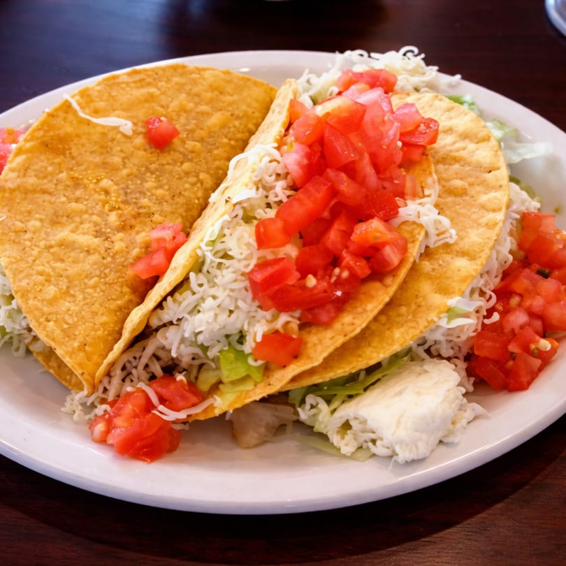Two crispy tacos filled with shredded lettuce, cheese, and diced tomatoes on a white plate.